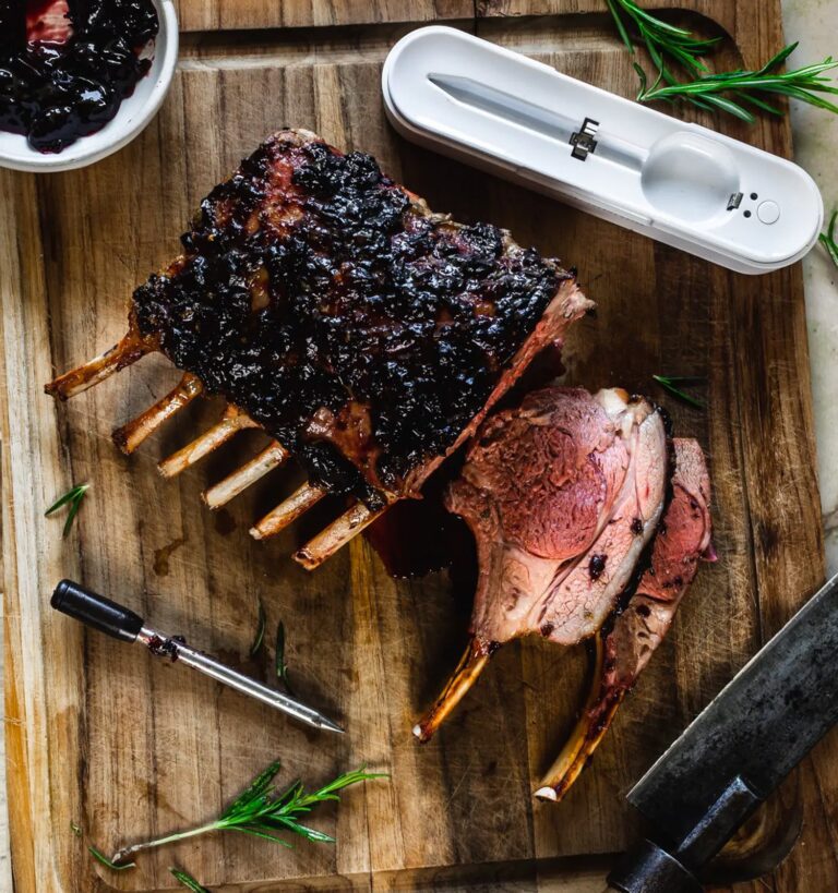 A cooked rack of lamb partially sliced on a wooden cutting board, garnished with herbs, with a meat thermometer, carving knife, and a small bowl of dark sauce nearby.