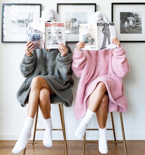 Two people in bathrobes with towels on their heads sit on stools, reading magazines, with framed photos on the wall behind them.