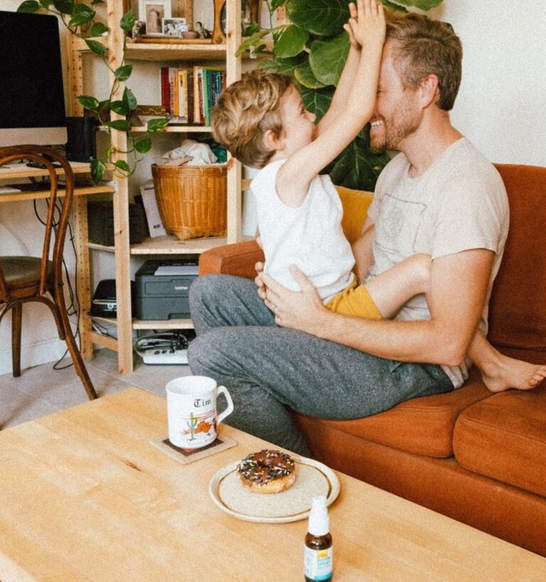 An adult and child sit on an orange couch, smiling and playing. A coffee mug, donut, and small spray bottle are on the wooden table in front of them in a cozy living room.