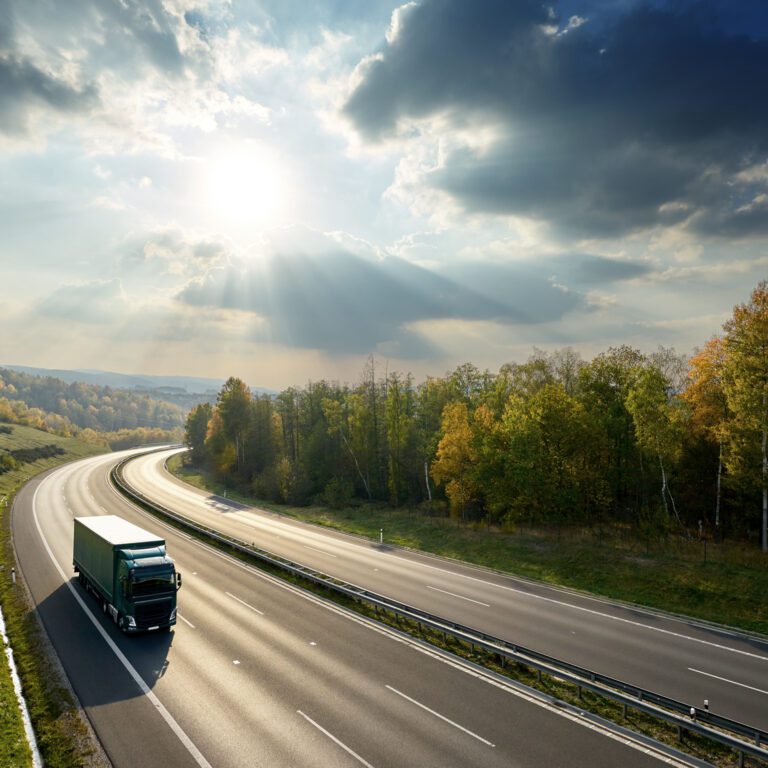 A green semi-truck drives on a curved, empty highway surrounded by trees under a partly cloudy sky with sunbeams.