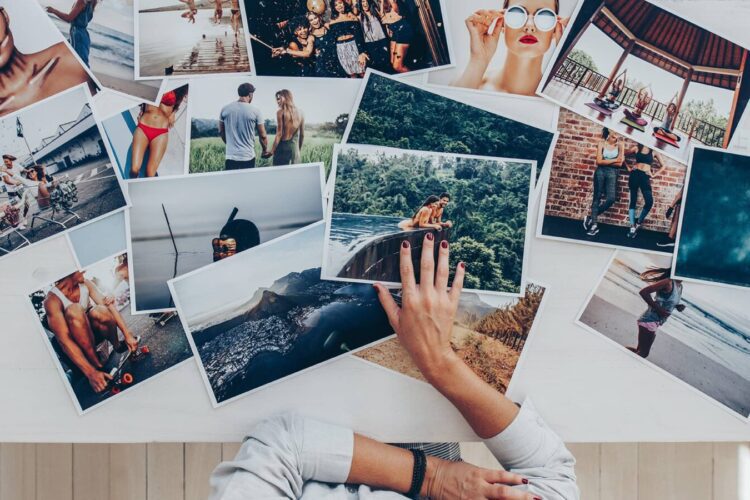 A person sits at a table covered with various photos, showing people in different outdoor and travel settings, including the beach, a forest, and urban scenes.