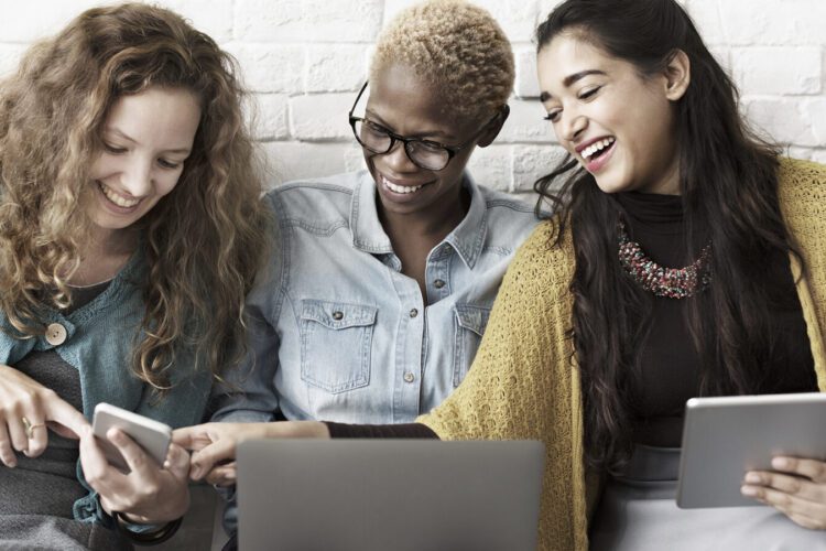 Three women sit together, smiling and looking at electronic devices including a smartphone, a laptop, and a tablet against a white brick wall.
