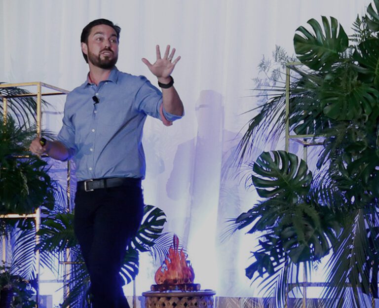 A man in a blue shirt speaks on stage with a microphone, gesturing with his left hand, surrounded by green plants and a white backdrop.