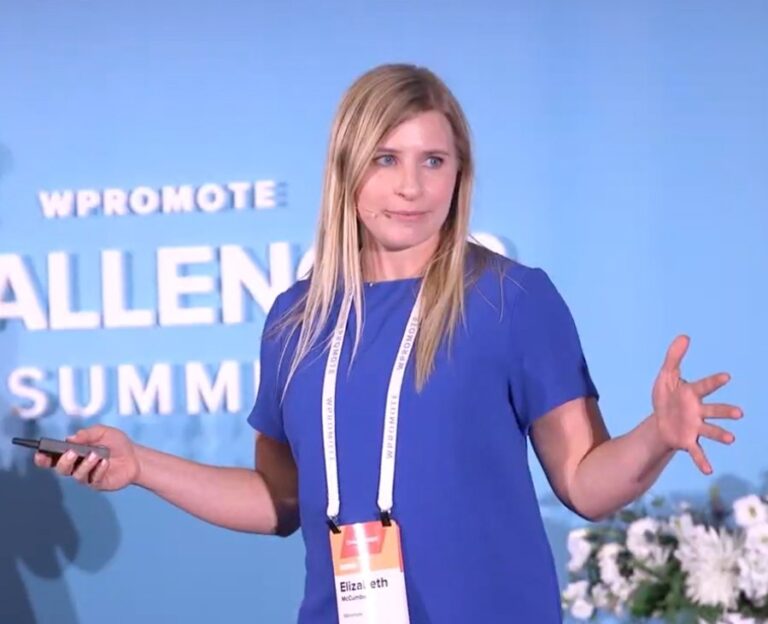 A woman in a blue dress speaks on stage at a summit, holding a remote and gesturing with her hands. She wears a badge labeled "Elizabeth.