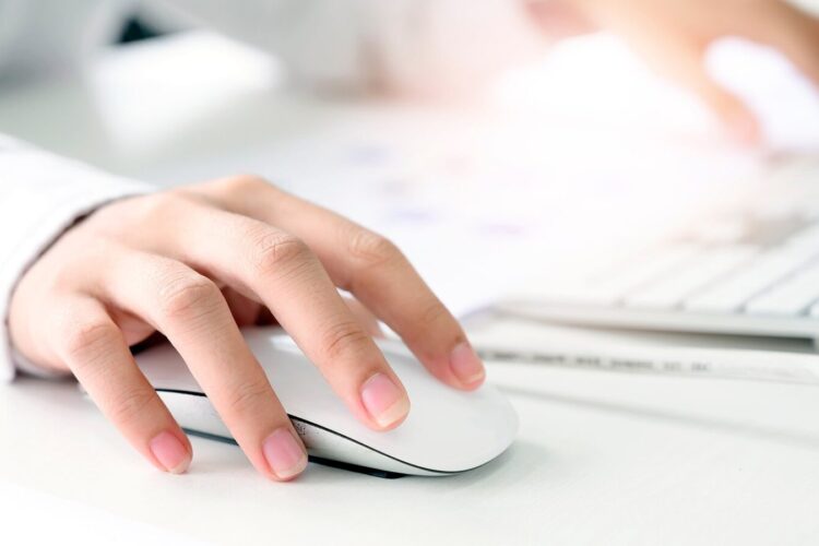 A person’s hand using a white computer mouse on a desk with another hand typing on a white keyboard in the background.