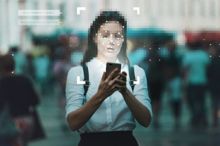 A person stands in a busy outdoor area, looking at a smartphone. Digital facial recognition graphics are overlaid on their face, indicating biometric scanning.