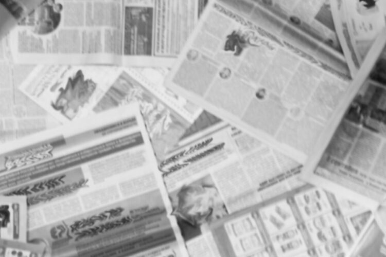 Several newspapers are spread out in an overlapping, scattered manner, with text and images visible but out of focus.