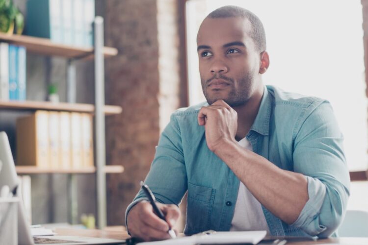 A man sits at a desk with a notebook and pen, resting his chin on his hand and looking thoughtfully into the distance.
