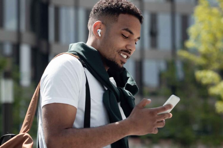 A man wearing wireless earbuds and a white t-shirt looks at his smartphone while standing outdoors, with a backpack over one shoulder.