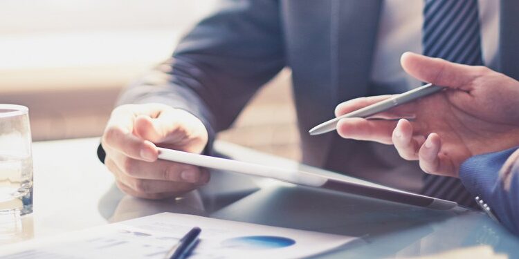 Two people in business attire review documents and a tablet at a glass-top desk with a laptop, papers, pen, and a glass of water.