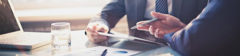 Two people in business attire review documents and a tablet at a glass-top desk with a laptop, papers, pen, and a glass of water.
