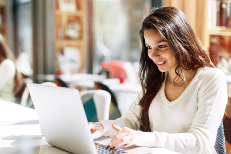 A woman with long brown hair sits at a table using a laptop, smiling, in a bright room with bookshelves and large windows.