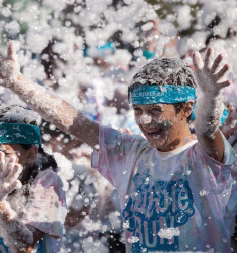 Children with headbands and t-shirts participate in a foam run event, smiling and raising their arms as they are covered in white foam.