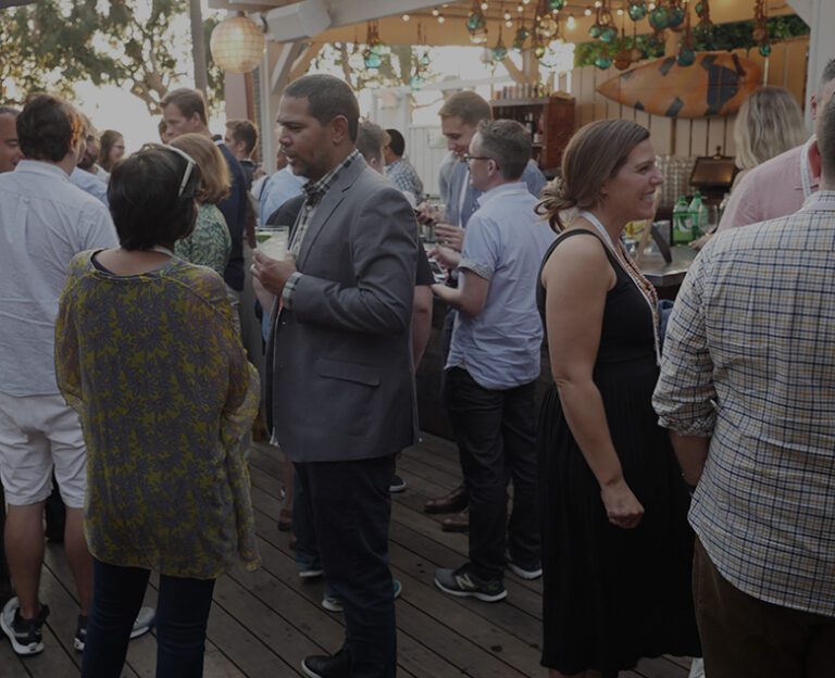 A group of people stand and converse at an outdoor social event, some holding drinks, on a wooden deck under string lights.