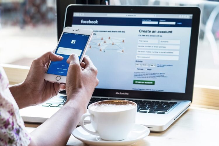 A person holding a smartphone with the Facebook login screen open, sitting at a table with a laptop displaying the Facebook sign-in page and a cup of coffee.