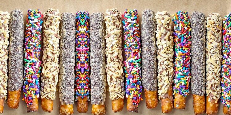 A row of pretzel rods coated with chocolate and decorated with colorful sprinkles and shredded coconut, arranged on a brown surface.
