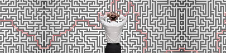 A person stands facing a large wall-sized maze with a red path highlighted, hands resting on the back of their head.