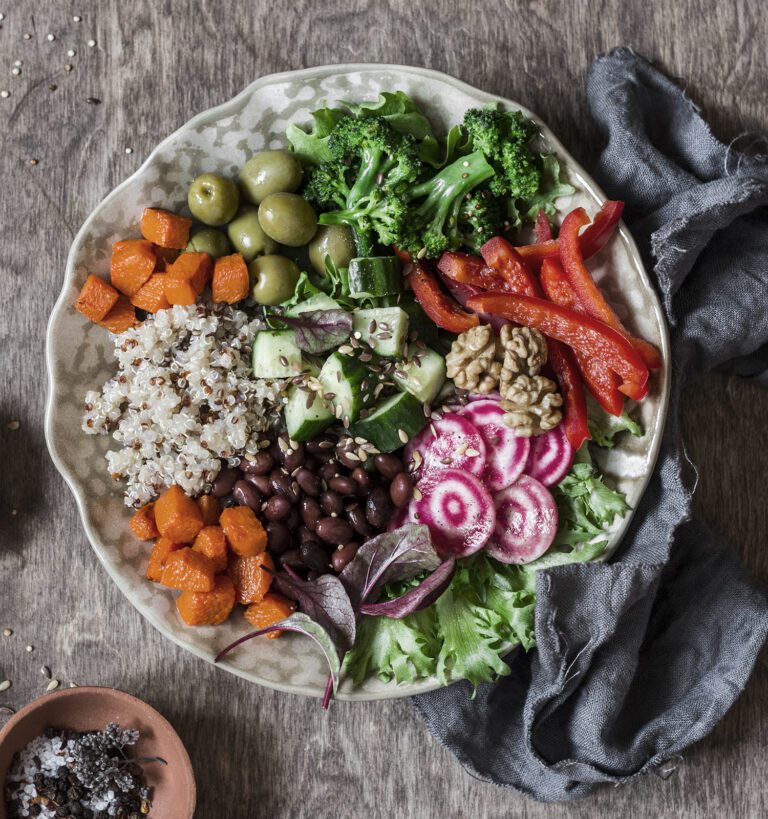A plate with quinoa, black beans, roasted sweet potatoes, broccoli, olives, cucumber, red bell pepper, walnuts, sliced radishes, and mixed greens on a wooden table.