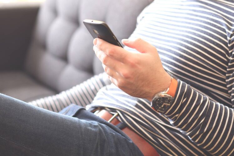 Person in a striped shirt sitting on a sofa, holding a smartphone in their right hand, wearing a wristwatch and jeans.