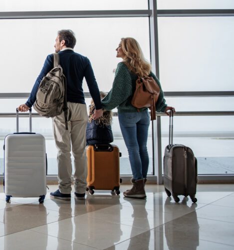 A man, woman, and child holding hands stand with luggage in an airport terminal, looking out large windows toward the runway.