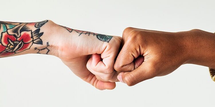 Two people doing a fist bump; one arm has tattoos and a plaid sleeve, the other has brown skin and a yellow striped sleeve, against a plain background.