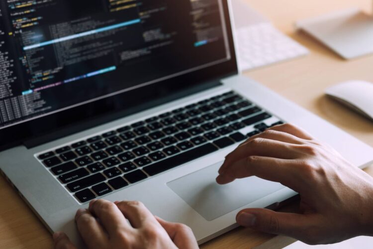 Person typing on a laptop with code displayed on the screen, placed on a wooden desk with a keyboard and mouse in the background.