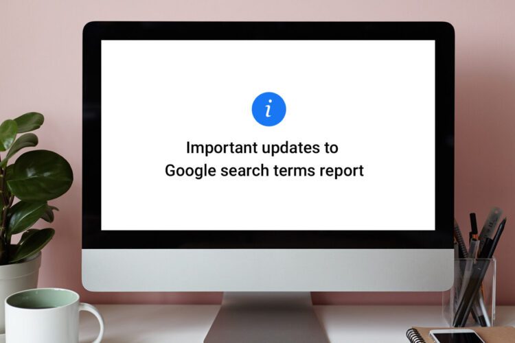 A computer monitor displays a message about important updates to the Google search terms report. A plant, pen, and notebook sit on the desk beside it.