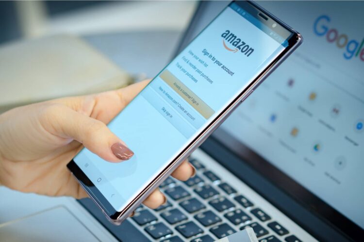 A person holds a smartphone showing the Amazon sign-in page, with a laptop displaying the Google homepage in the background.