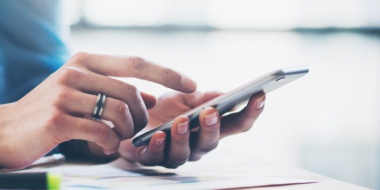 Close-up of a person using a smartphone at a desk, with papers and pens visible in the background.
