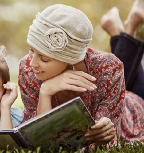 A woman wearing a headscarf lies on the grass, reading a book to a child beside her.