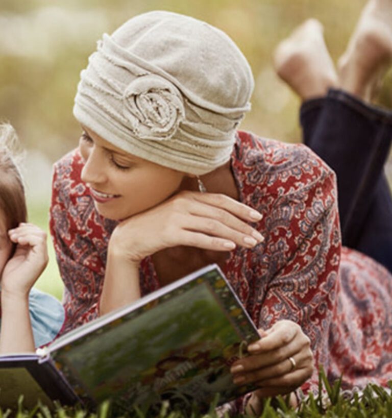 A woman wearing a headscarf lies on the grass, reading a book to a child beside her.