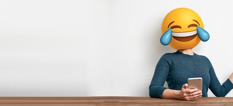 Person sitting at a table holding a smartphone with a large laughing-with-tears emoji covering their face.