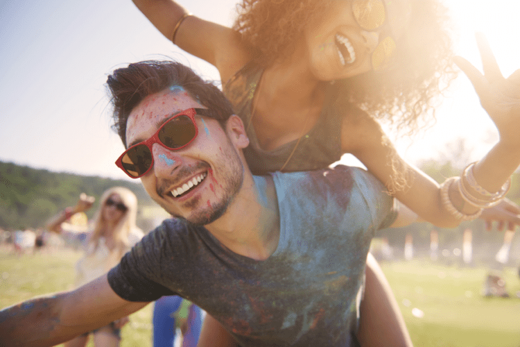 Two people with painted faces and sunglasses smile as one gives the other a piggyback ride outdoors at a festival.