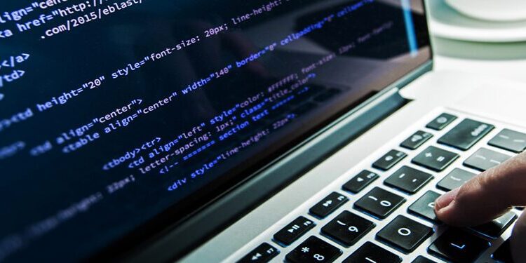 Close-up of a person typing on a laptop keyboard with lines of code displayed on the screen; a smartphone and coffee cup are visible in the background.