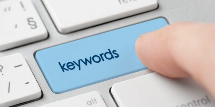 A finger presses a blue keyboard key labeled "keywords" on a silver laptop keyboard.