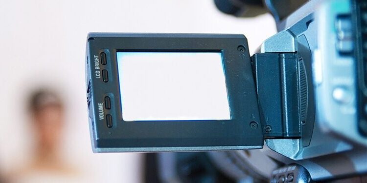 A video camera with a flip-out screen records two people, likely a bride and groom, standing together in the blurred background.