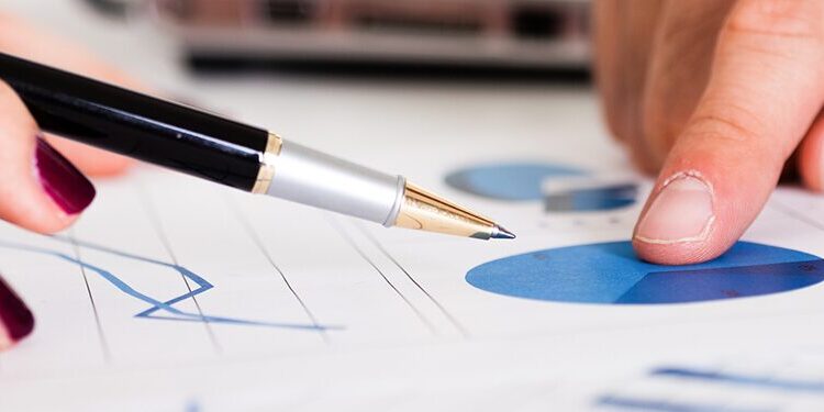 Two people review documents with blue charts and graphs; one points at a pie chart while the other holds a pen above a line graph.