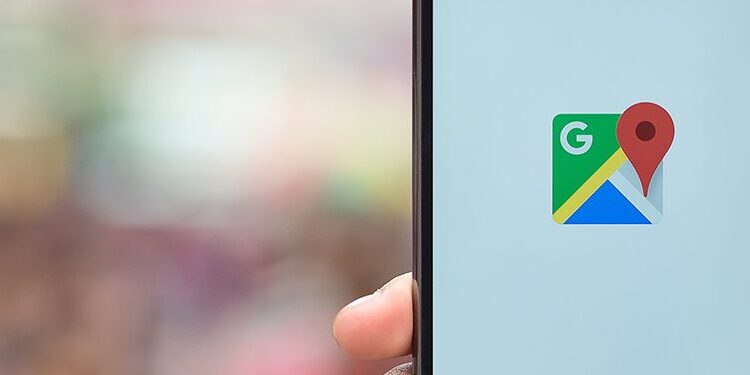 A hand holds a smartphone displaying the Google Maps app logo on the screen, with a blurred background.