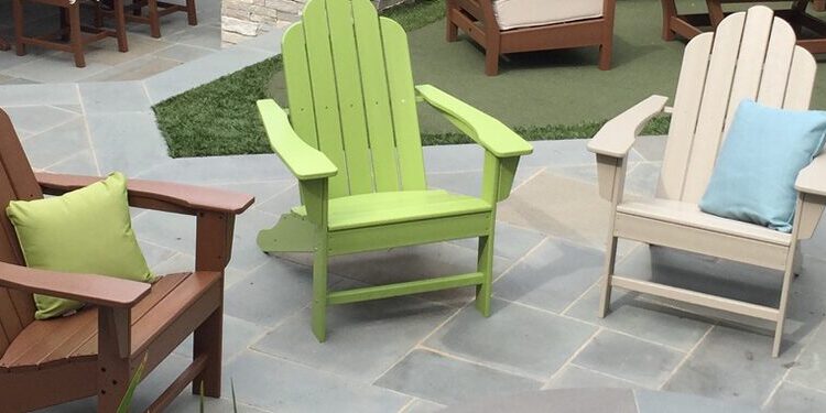 Four Adirondack chairs in brown, green, white, and blue with cushions are arranged in a circle on a stone patio, with outdoor seating sets visible in the background.