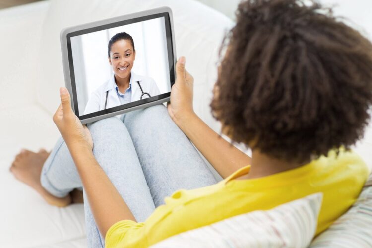 A person sitting on a couch holds a tablet, participating in a video call with a doctor who is smiling on the screen.