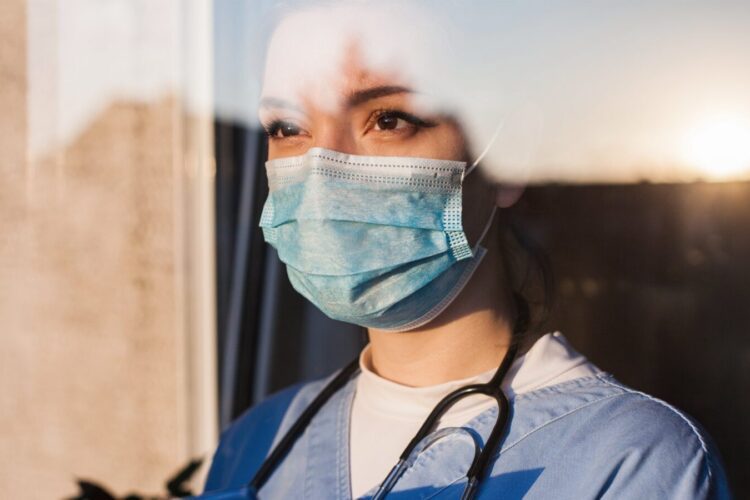A healthcare worker in scrubs, wearing a face mask and stethoscope, looks out a window with sunlight in the background.