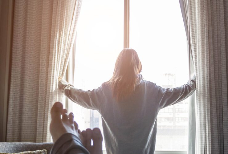 Person standing at a window opening curtains with sunlight coming in, seen from the perspective of someone lying on a bed.