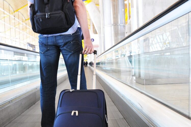 Person with a backpack and rolling suitcase walks on a moving walkway in an airport terminal.