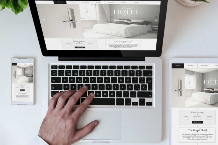 A person using a laptop with a hotel website displayed; the same website appears on a smartphone and tablet next to the laptop on a white desk.