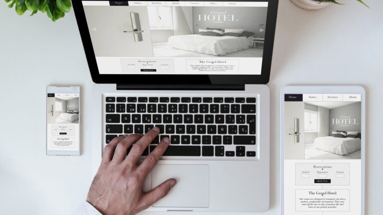 A person using a laptop with a hotel website displayed; the same website appears on a smartphone and tablet next to the laptop on a white desk.