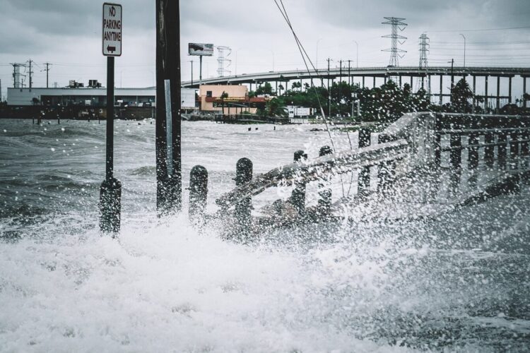 Large waves crash over a flooded roadway near a "No Parking" sign, with industrial buildings and power lines visible in the background under a cloudy sky.