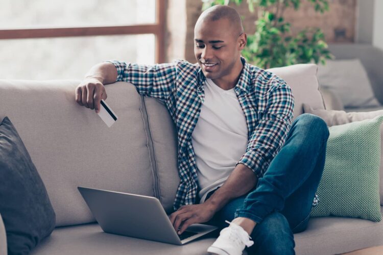 A man sits on a couch using a laptop and holding a credit card, appearing to make an online purchase.