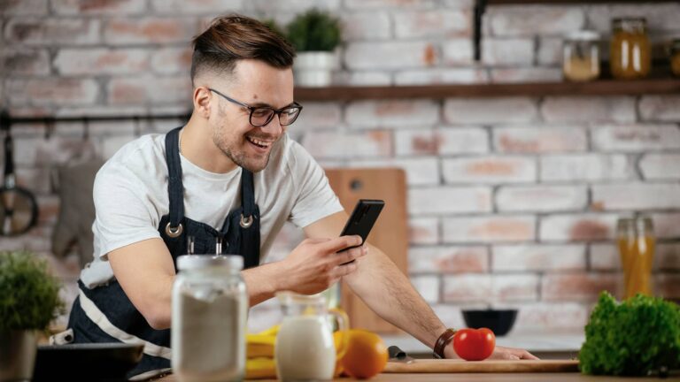 A person wearing an apron smiles while looking at a smartphone in a kitchen, with various ingredients and cooking utensils on the counter.