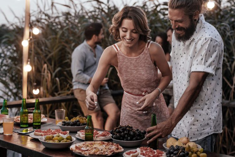 Two people serve themselves food from a table set with various dishes and drinks at an outdoor gathering, with others talking in the background.