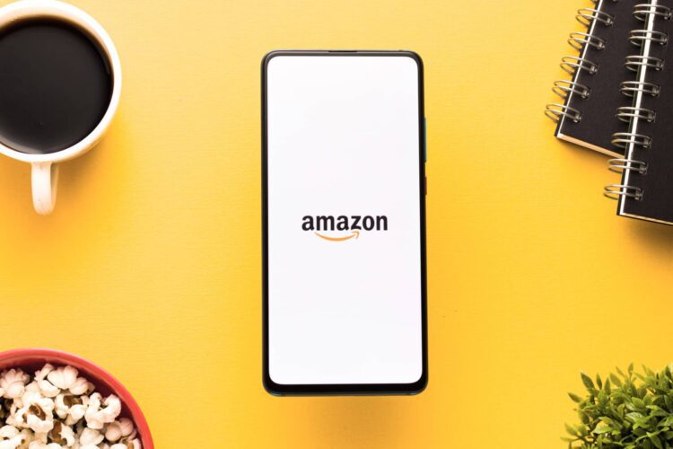 A smartphone displaying the Amazon logo is placed on a yellow surface, surrounded by a cup of coffee, notebooks, a bowl of popcorn, and a small green plant.